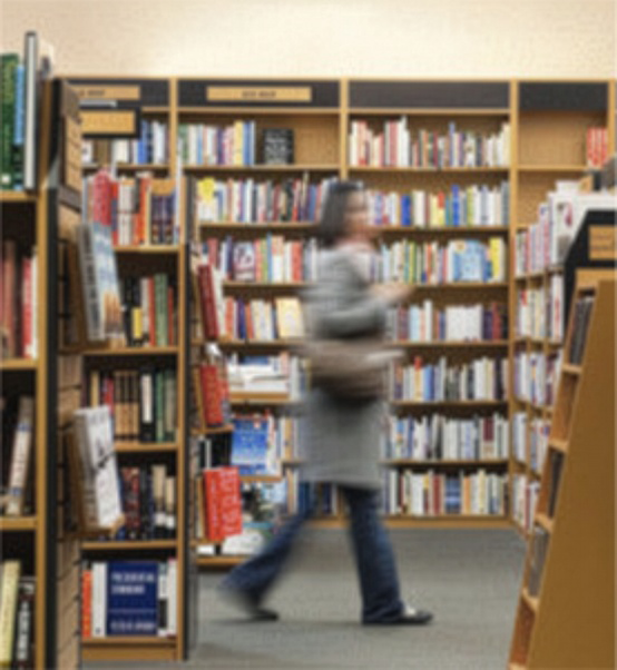 a cozy view of the bookshop with rare signed first editions in a lit bookcase to the right. To the left of a window showing a view of the New York City skyline, there is a bookcase filled with finely bound books.